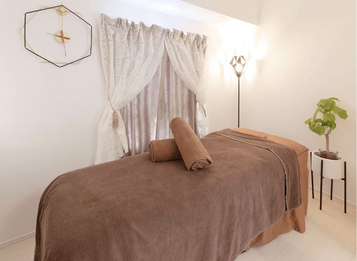 Massage table in a bright spa room with brown blanket, two rolled towels, sheer curtains, and a plant.