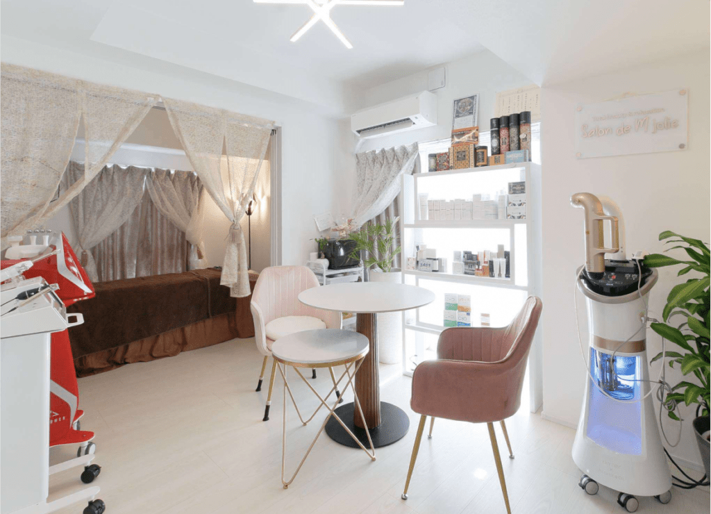 Bright beauty salon lounge with round table two pink chairs product shelves treatment bed behind sheer curtains and a skincare machine on the right